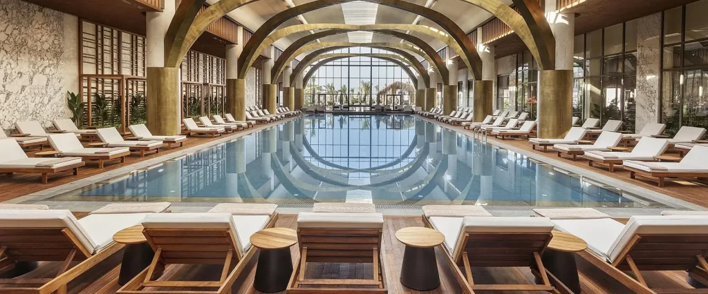 Cullinan Belek — indoor pool hall with brass arches and sun loungers, hotel interior design by ARTMİM